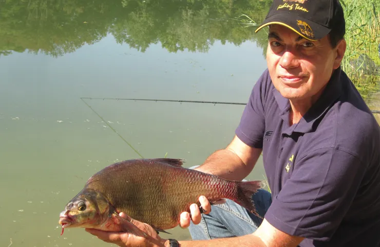 Angler steht an einem Teich und hält einen Süßwasserfisch in den Händen.